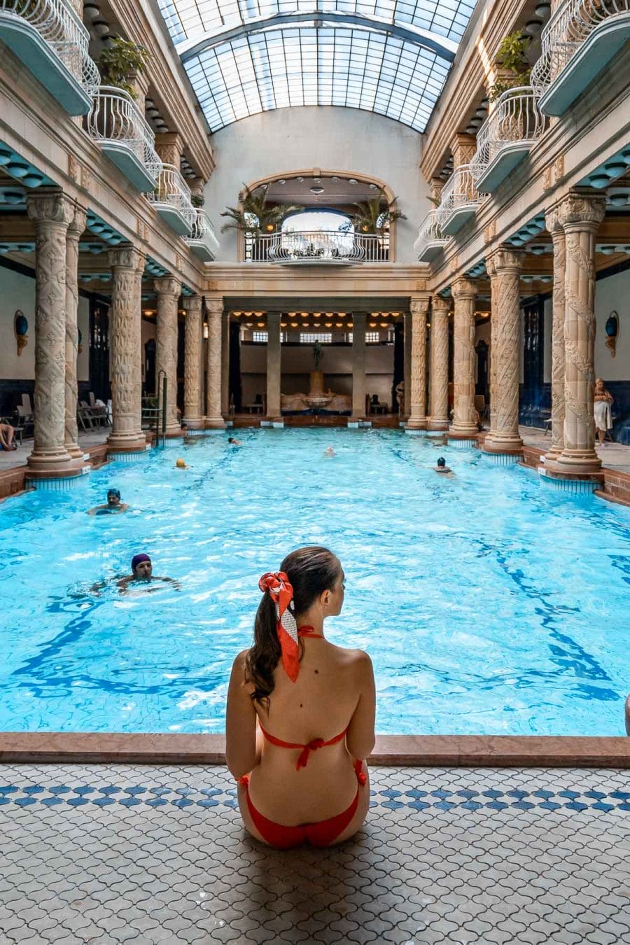 Girl in a red bikini sitting in front of a pool at the Gellert Baths in Budapest