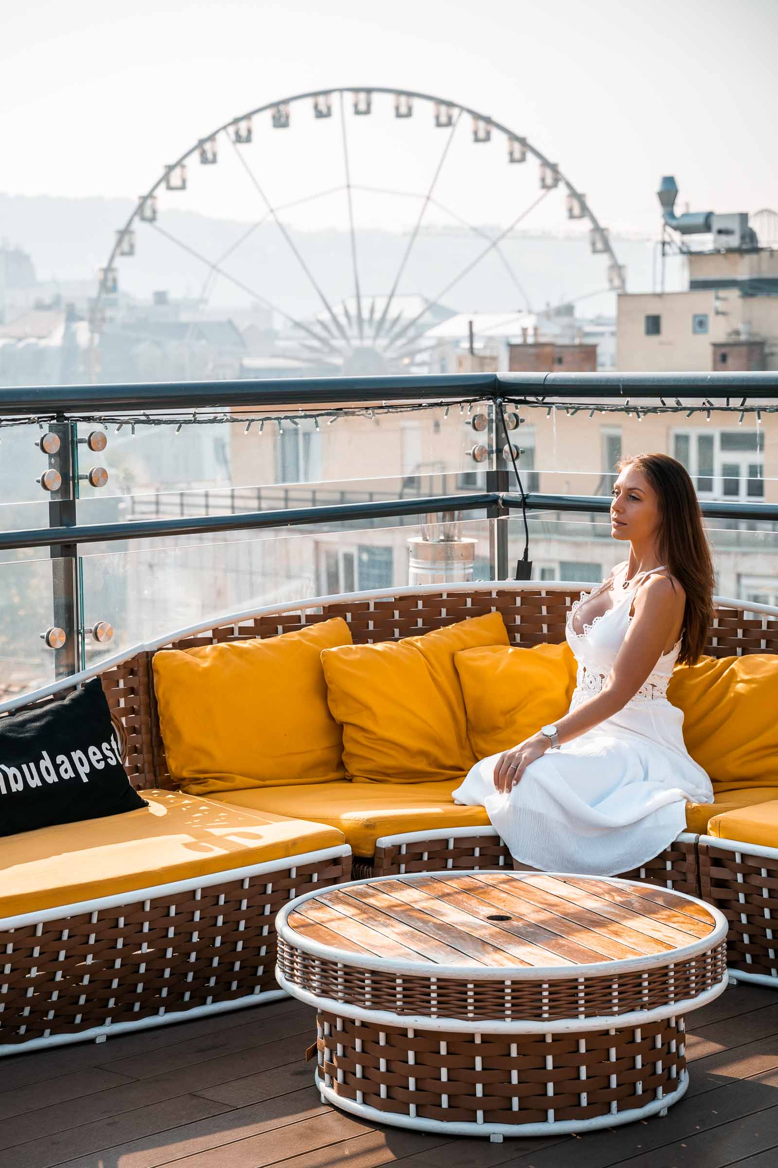 Girl in a white dress sitting in a yellow sofa at the High Note Skybar with the Budapest Eye in the background