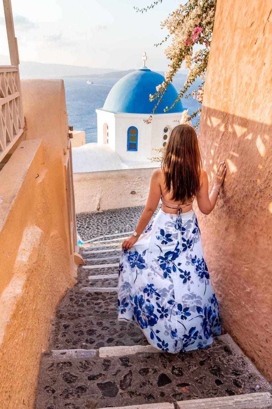 Girl in a blue floral dress walking down the stairs with a blue dome in front of her in Oia, Santorini