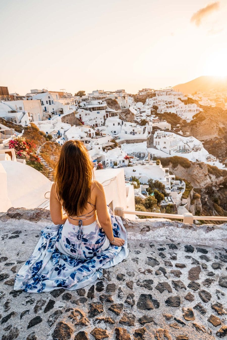 Girl in a blue floral dress sitting on a stone pathway, watching the sunrise in Oia, Santorini
