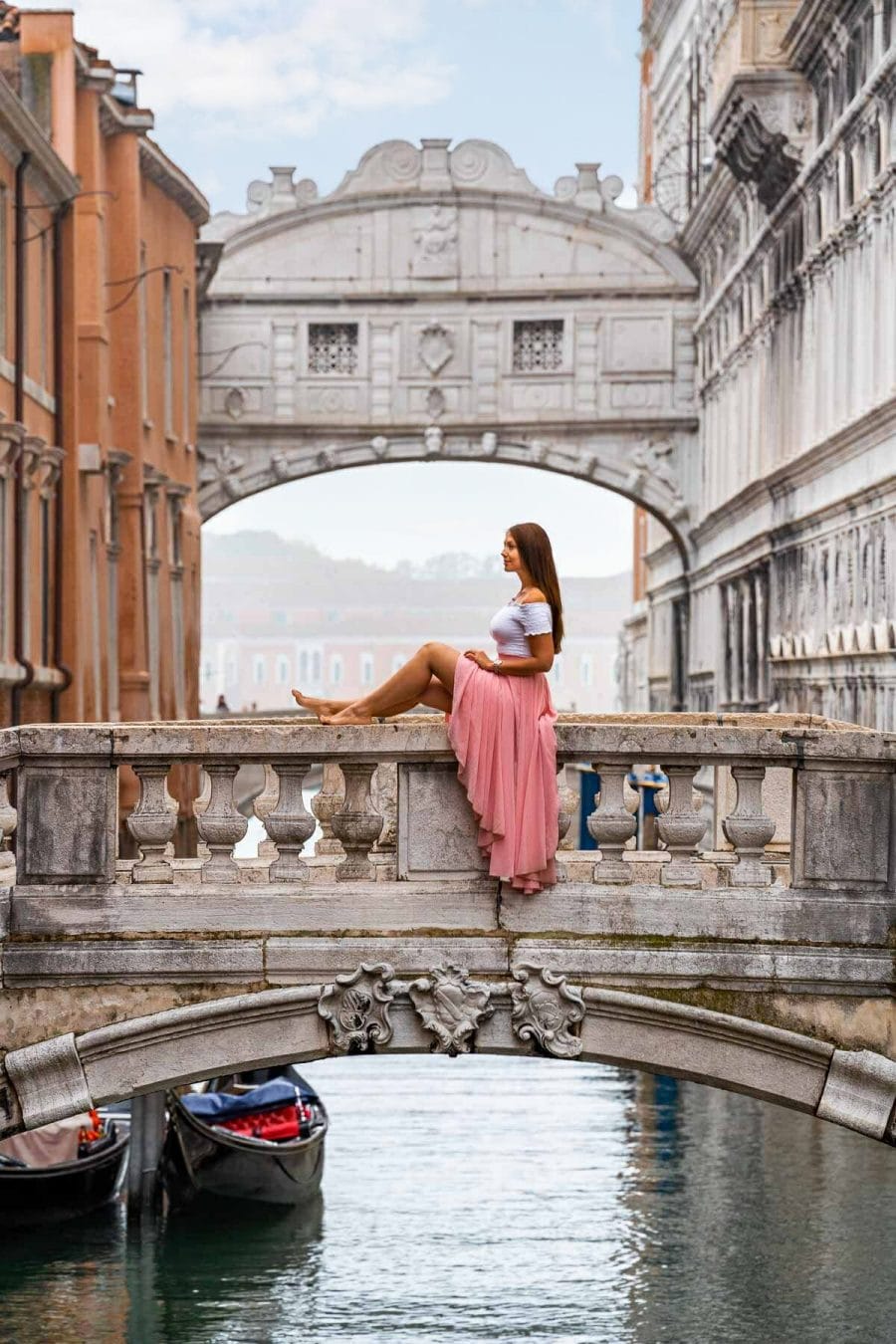 Girl in a pink skirt sitting on a stone bridge in front of the Bridge of Sighs in Venice