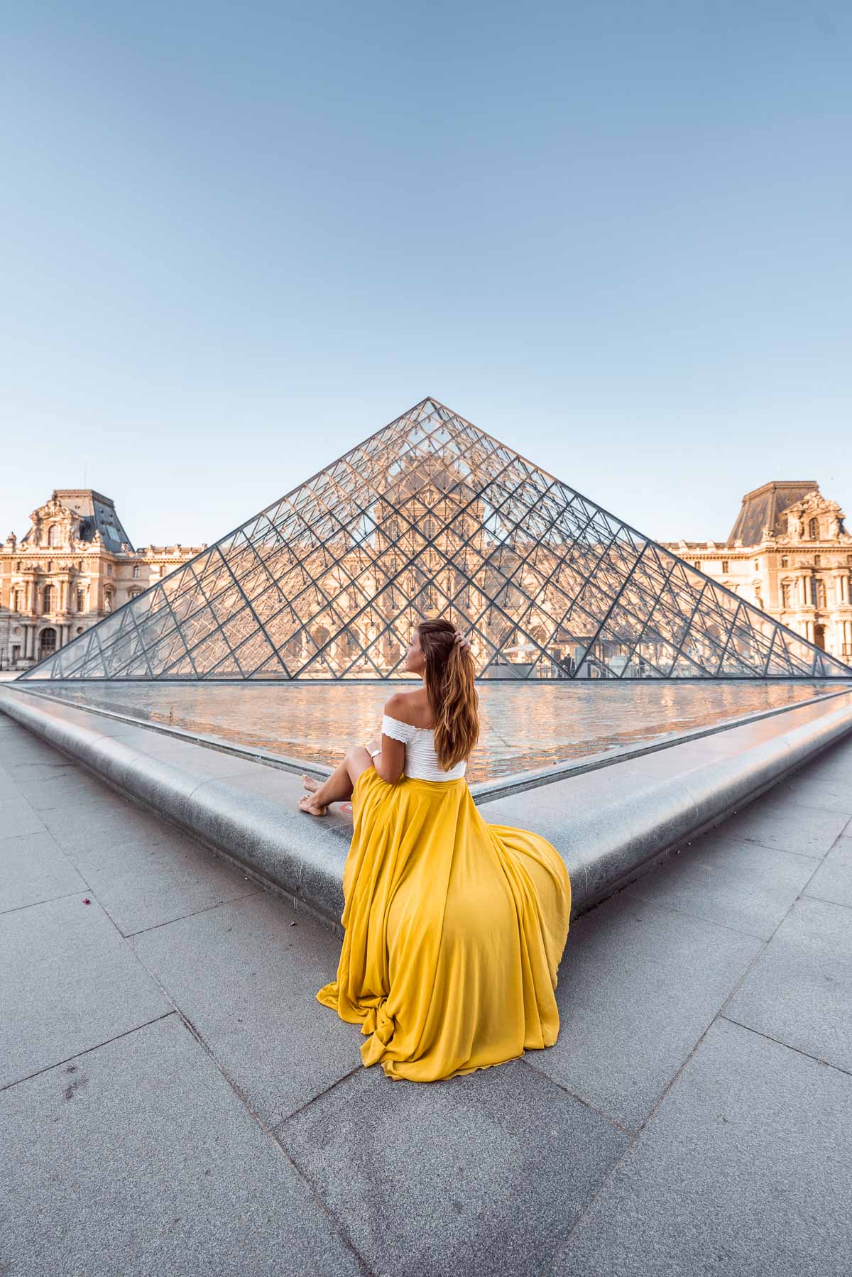 Girl in a yellow skirt sitting in front of one of the pyramids at the Louvre, one of the most instagrammable places in Paris
