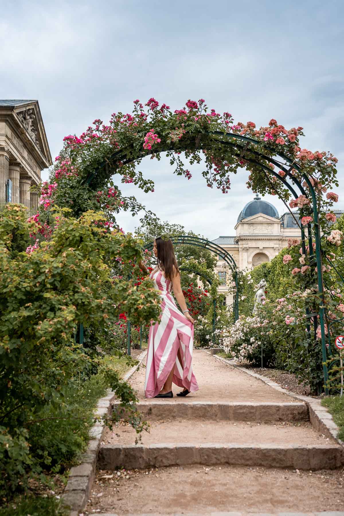 Girl in a pink-white striped dress standing under an archway of roses at Jardin des Plantes in Paris, France