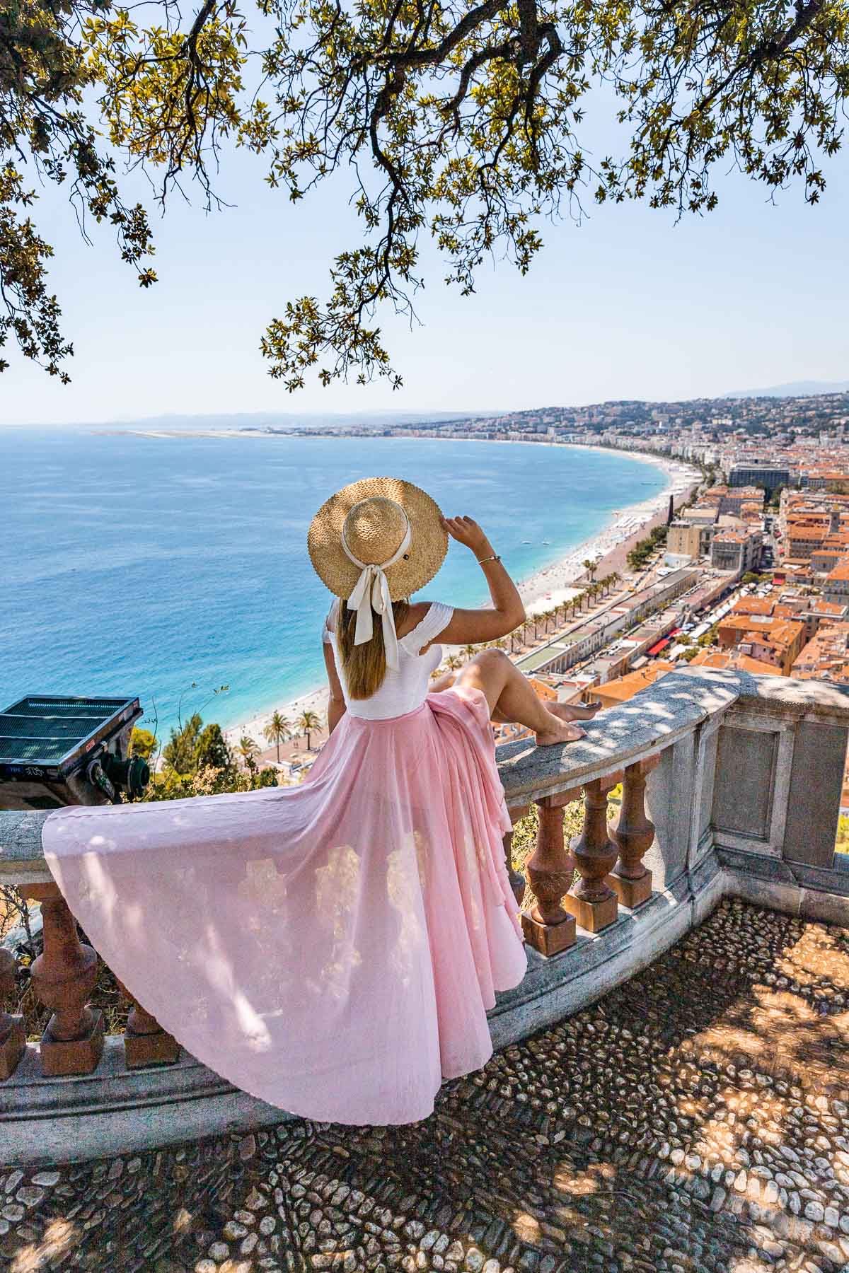 Girl in a pink skirt sitting on the railings at Castle Hill, looking at the panoramic view of Nice