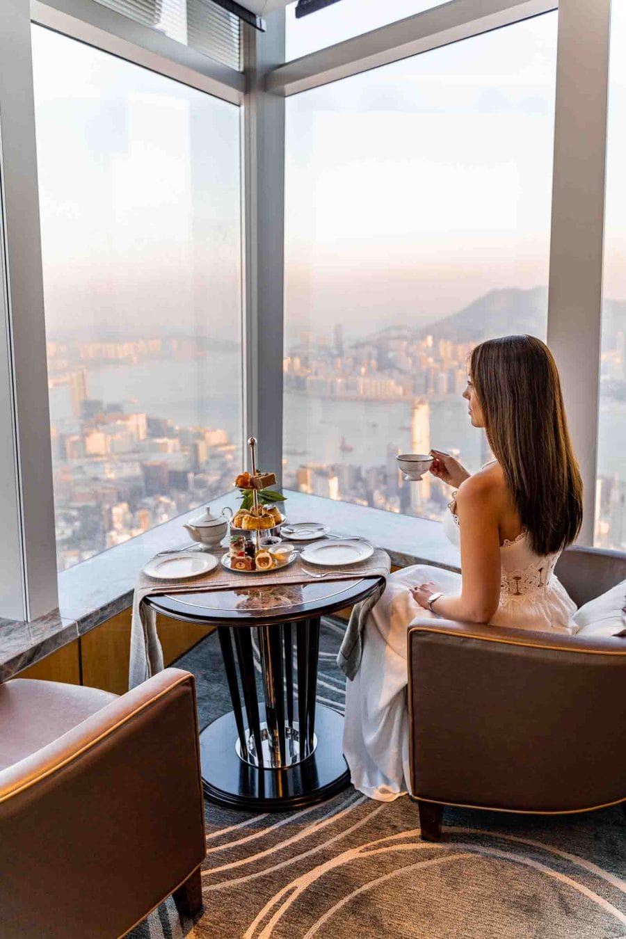 Girl in white dress drinking tea in front of a window with the view of the Hong Kong skyline