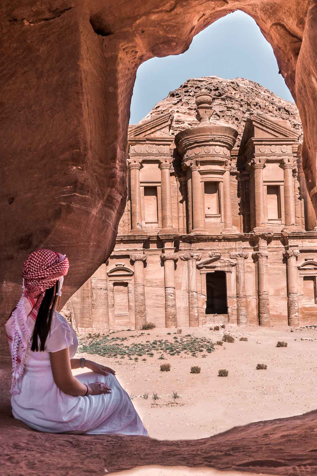 Girl in a white dress sitting in a cave, looking at the Al Deir Monastery in Petra, Jordan