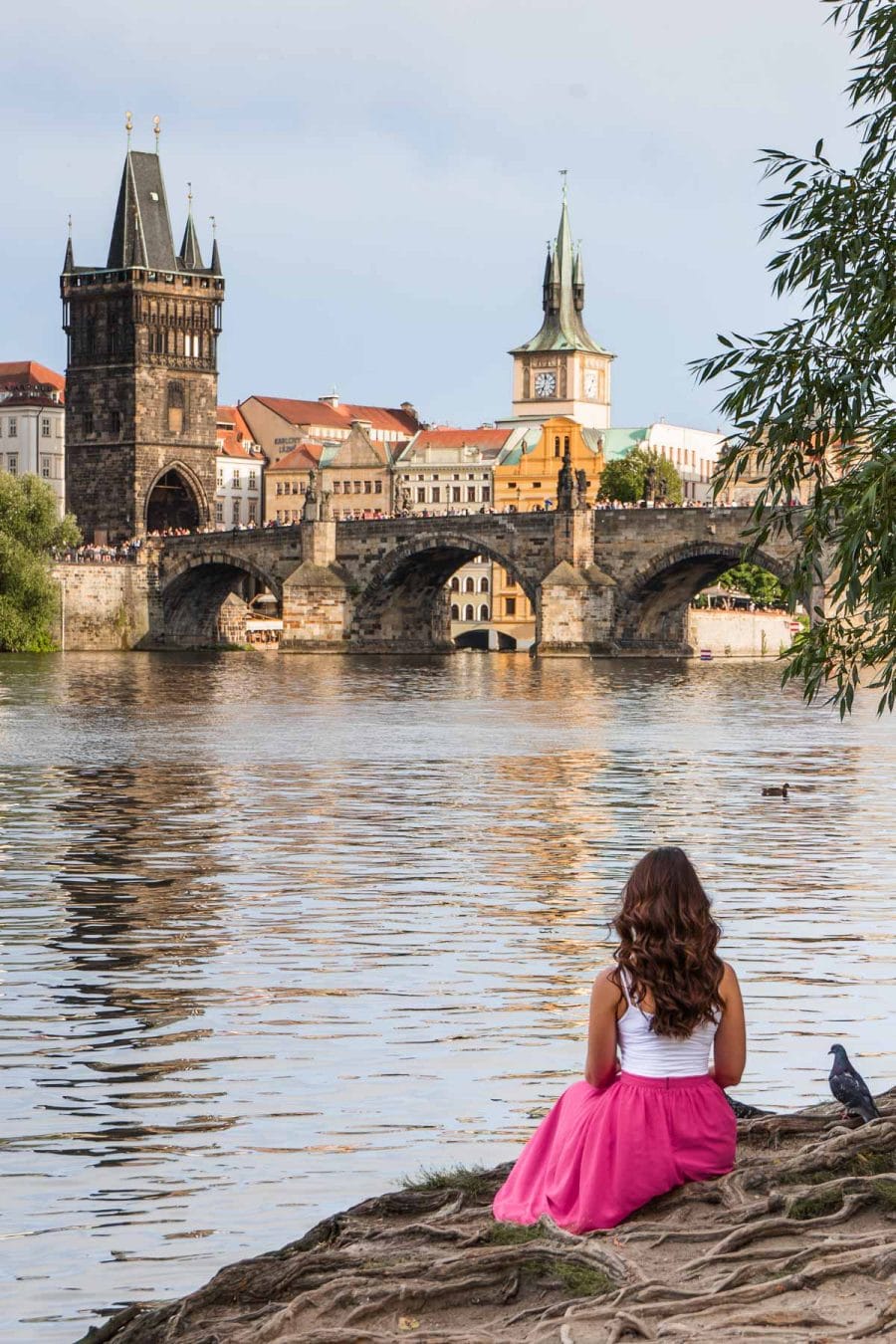 Girl in a pink skirt sitting by the river in Prague with the Charles Bridge in the background