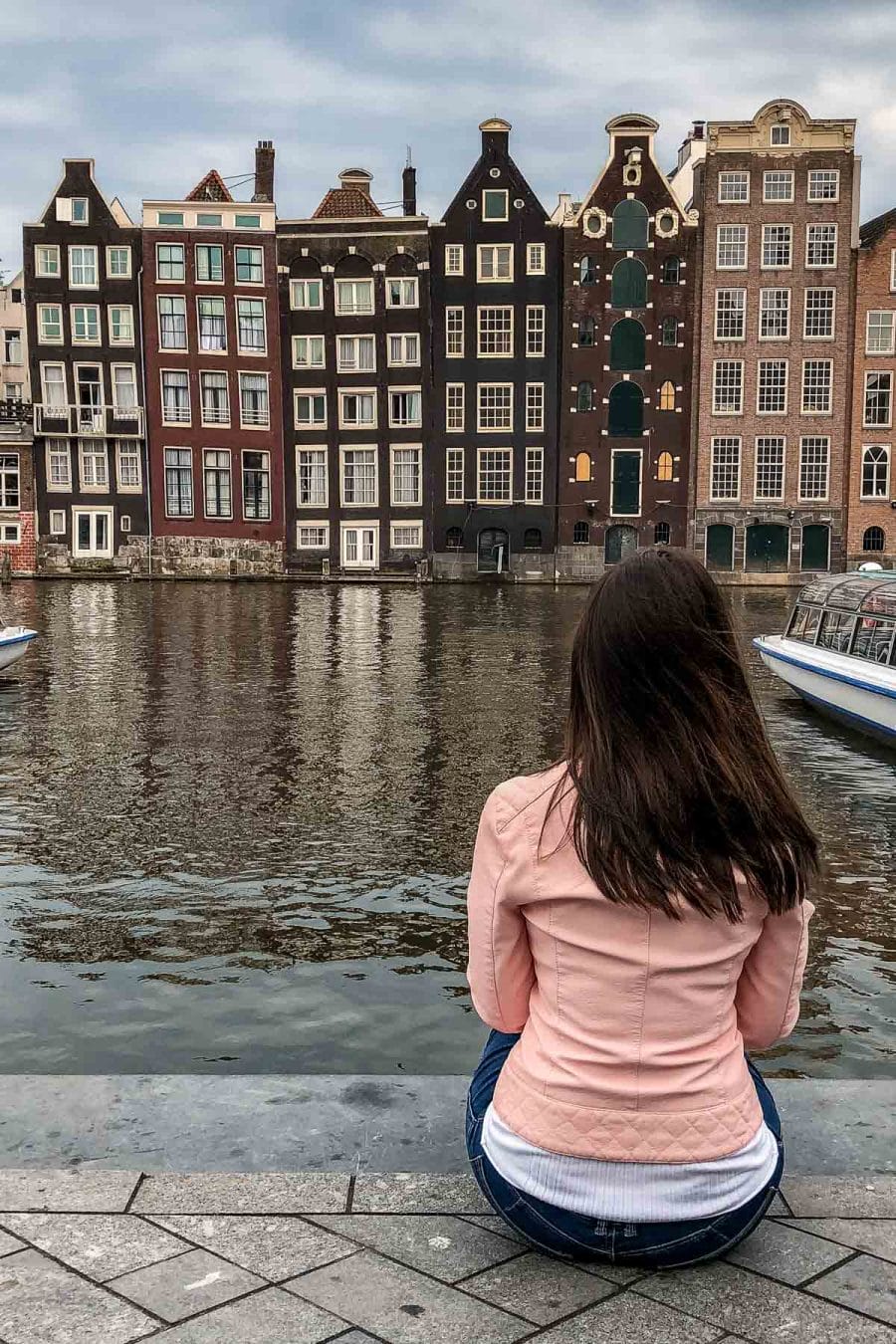 Girl in a pink jacket sitting in front of the crooked houses in Amsterdam