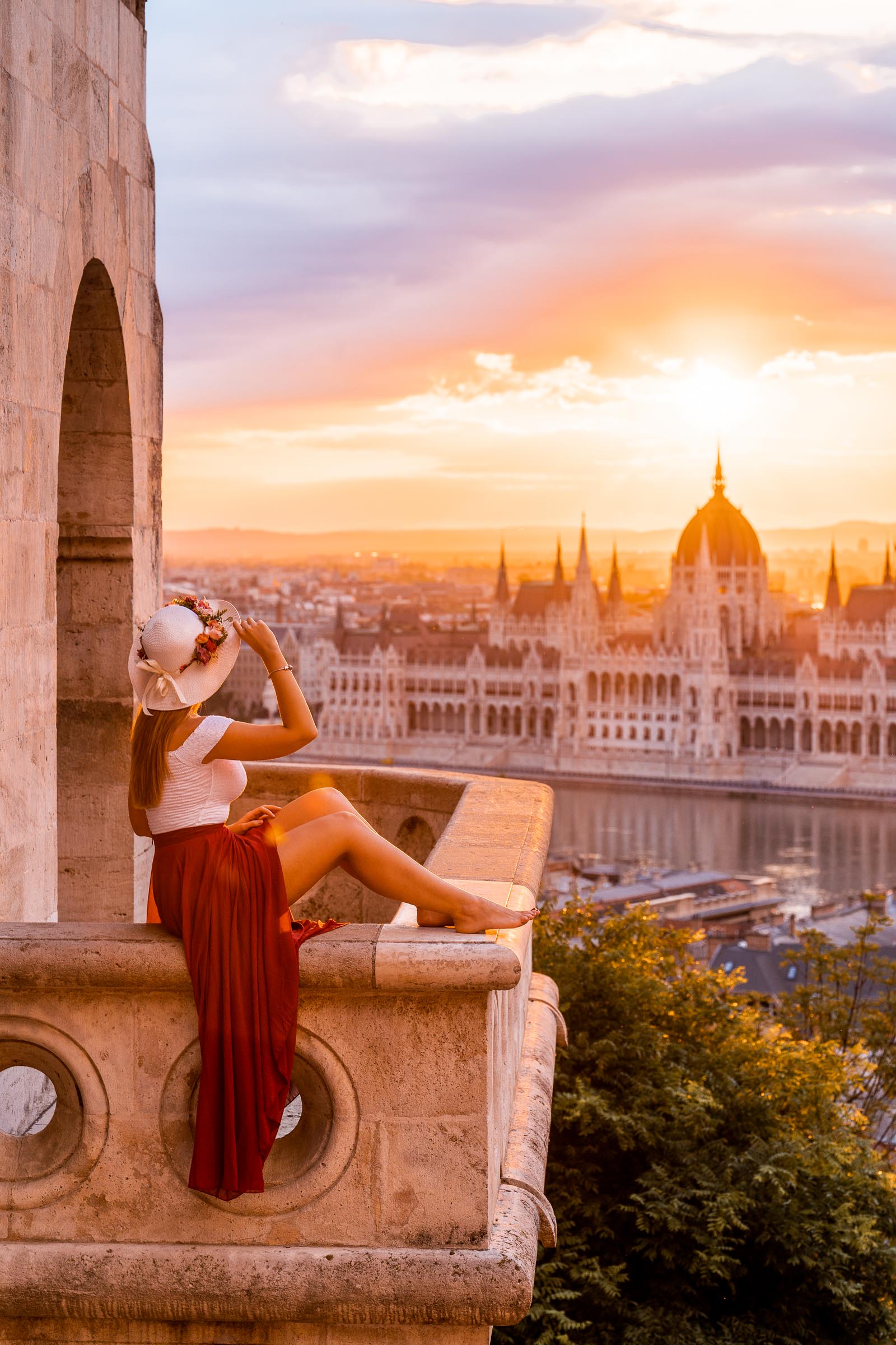 Girl in a red dress sitting on a balcony at the Fisherman's Bastion, looking at the Hungarian Parliament at sunrise