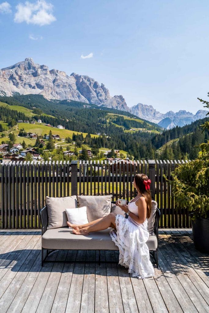 Girl in a white dress sitting on a sofa, looking at the mountain panorama at Hotel La Majun in the Dolomites