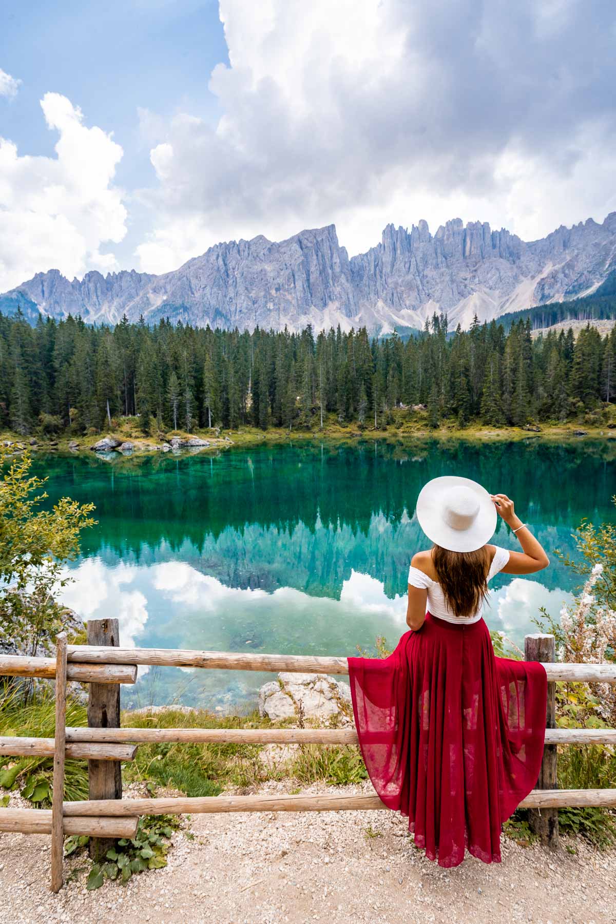 Girl in a red skirt sitting on the fence at Lago di Carezza, which is a must stop on every Dolomites road trip