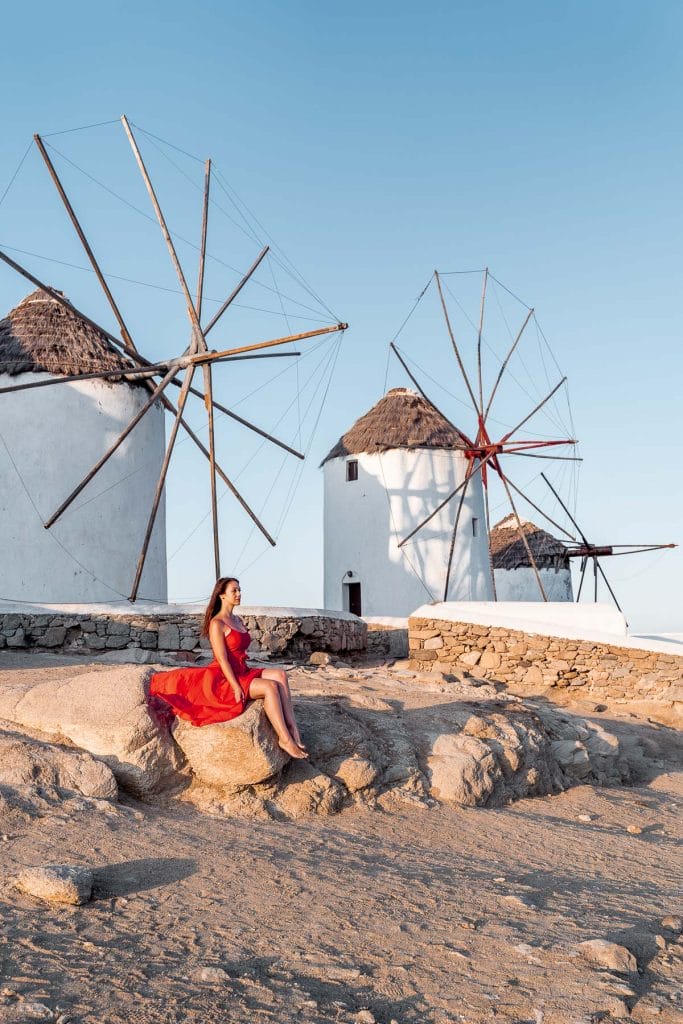 Girl in a red dress sitting on a rock in front of the windmills in Mykonos
