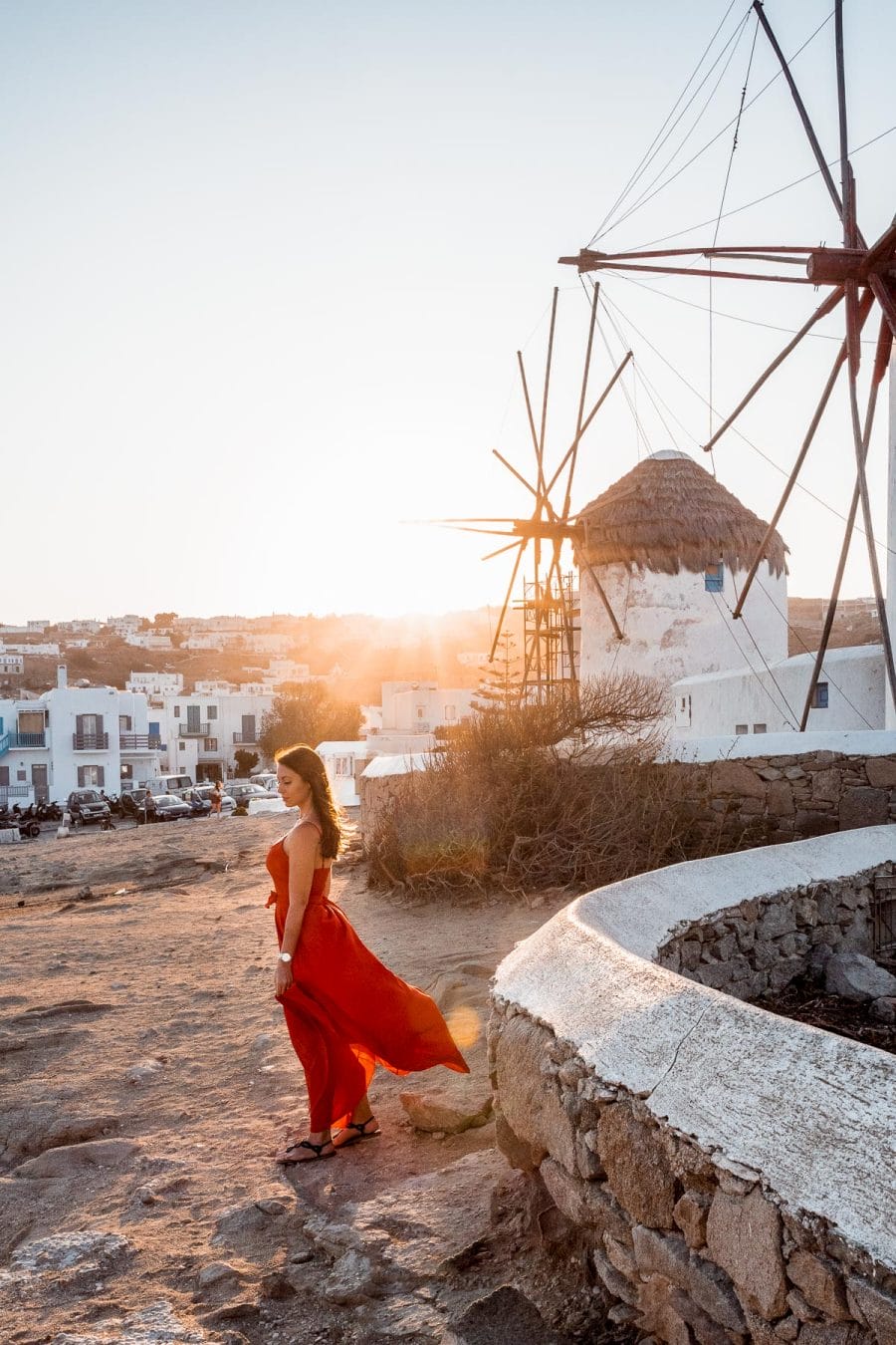 Girl in a red dress standing in front of the windmills in Mykonos at sunrise
