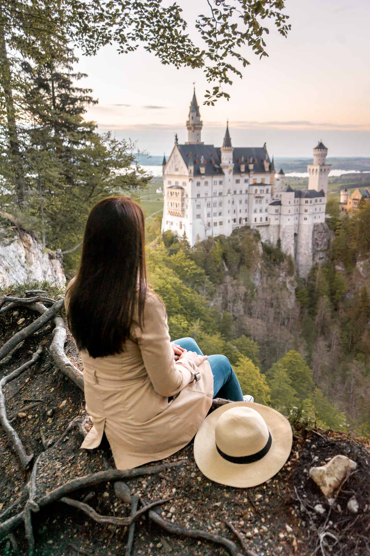 Girl in a trench coat sitting at the edge of a cliff, looking at the sunset over the Neuschwanstein Castle