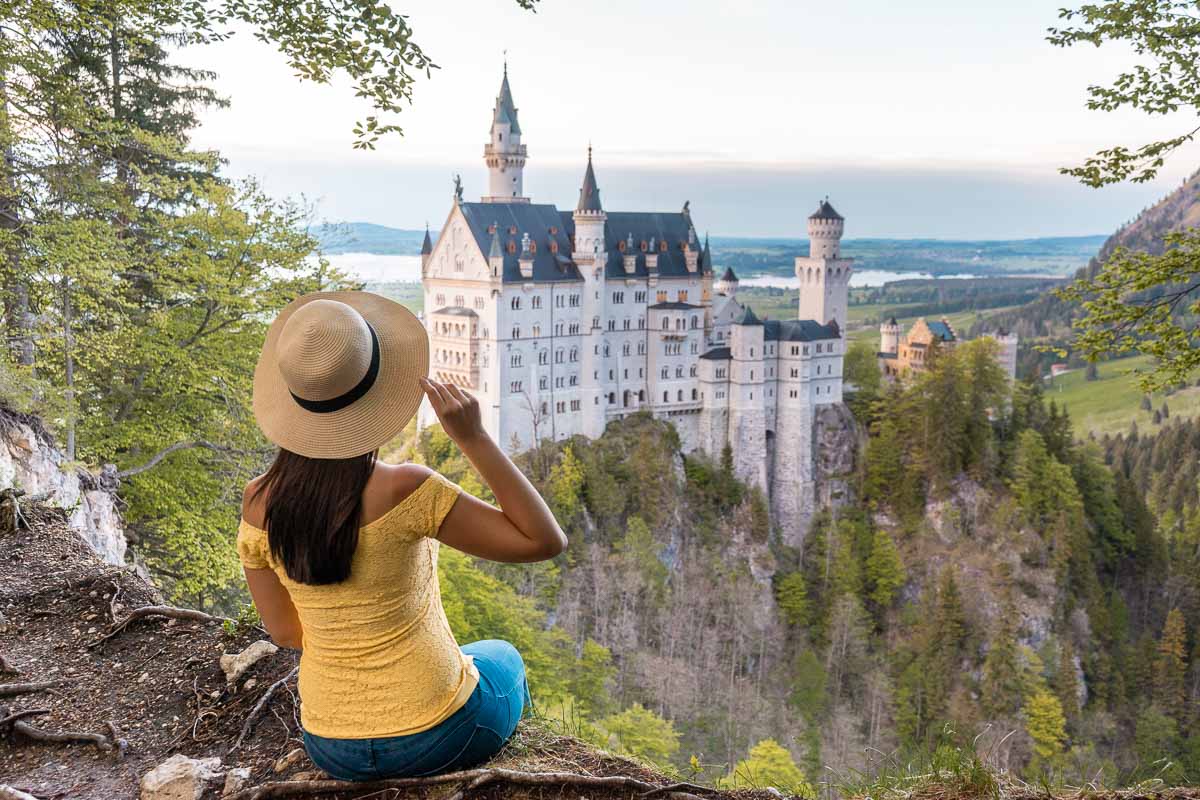 Girl in yellow top and a straw hat sitting at the edge of a cliff, looking at the sunset over the Neuschwanstein Castle