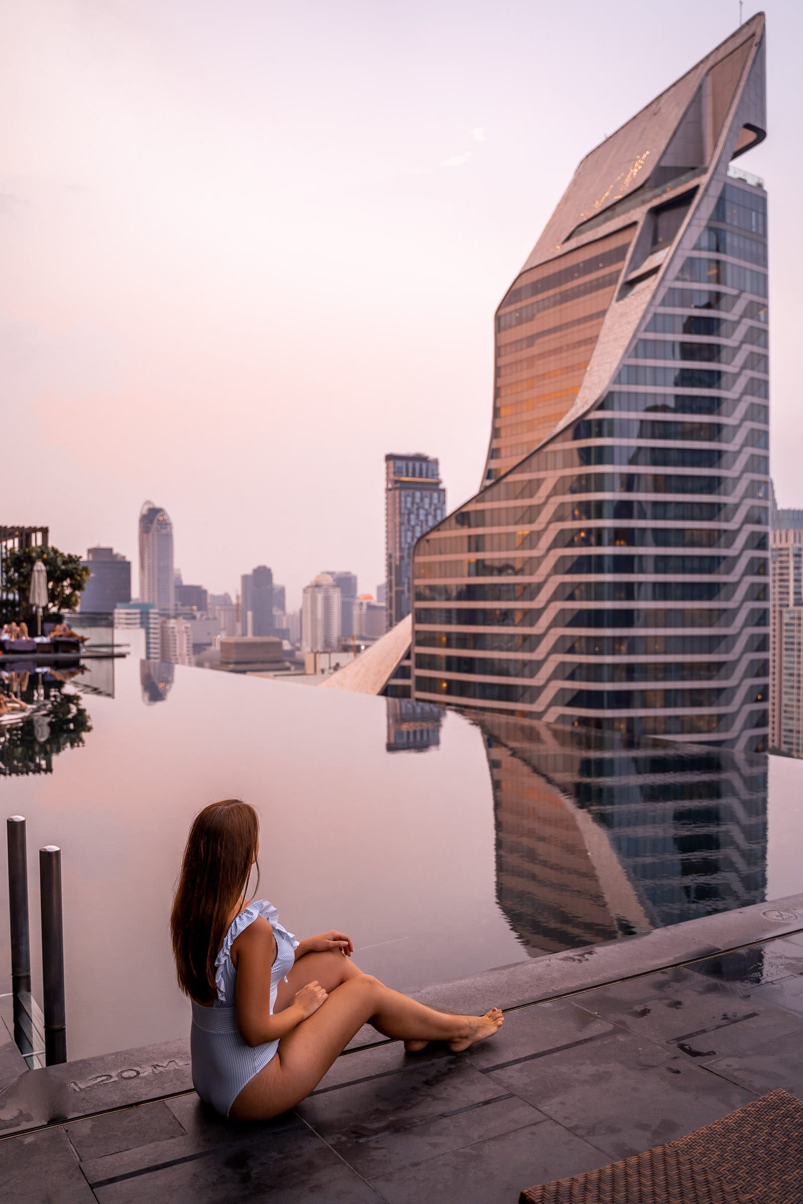 Girl in a blue swimsuit sitting in front of the infinity pool at the Okura Prestige Bangkok watching the sunset with the Bangkok skyline in the background