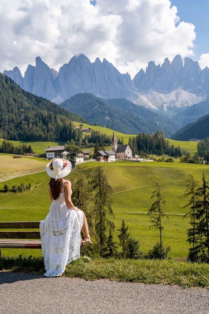 Girl in a white dress sitting on a bench, looking at Santa Maddalena church in Val di Funes, which is a most visit on a Dolomites road trip