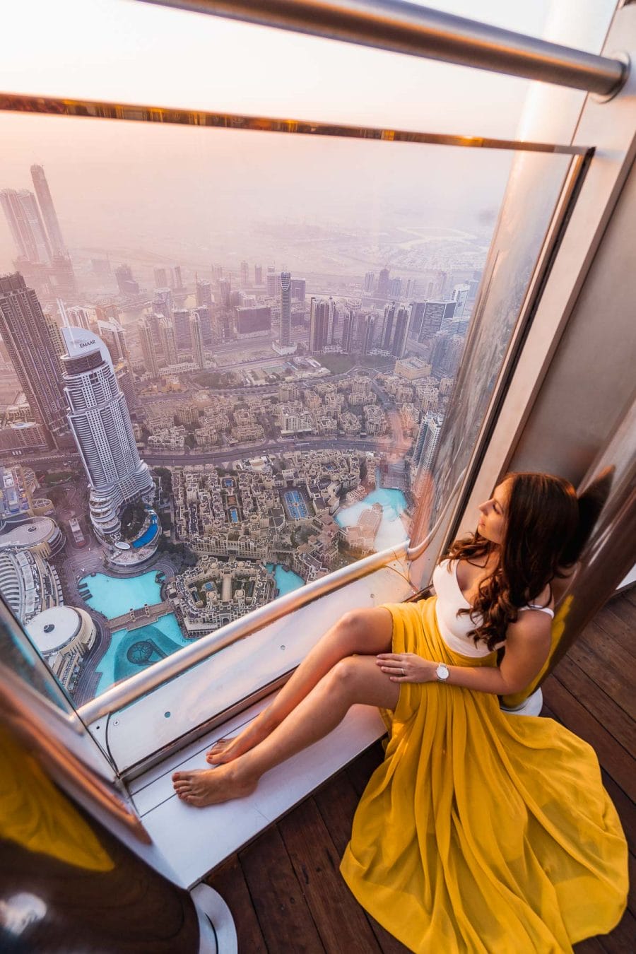 Girl in a yellow skirt sitting in front of a window in the Burj Khalifa at sunrise with the Dubai skyline in the background