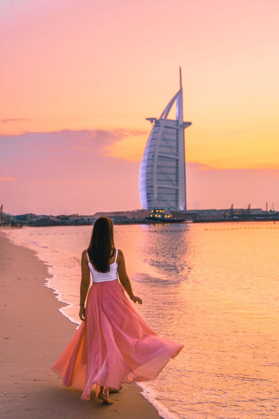 Girl in a flowy pink dress walking at the Jumeirah Beach at sunset with the Burj al Arab in the background