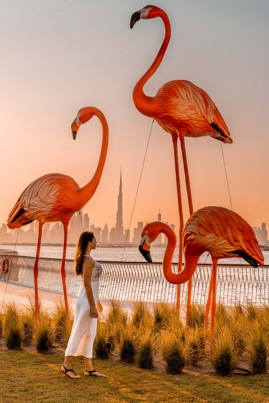 Girl in a white dress standing in front of pink flamingos at the Dubai Creek Harbour at sunset
