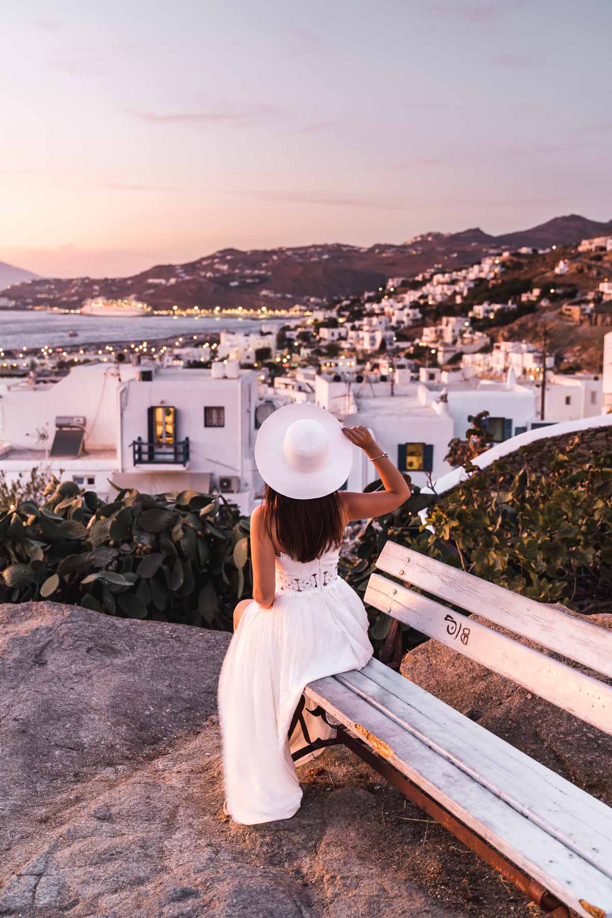 Girl in a white dress sitting on a bench at Boni's Windmill, watching the sunset over Mykonos Town