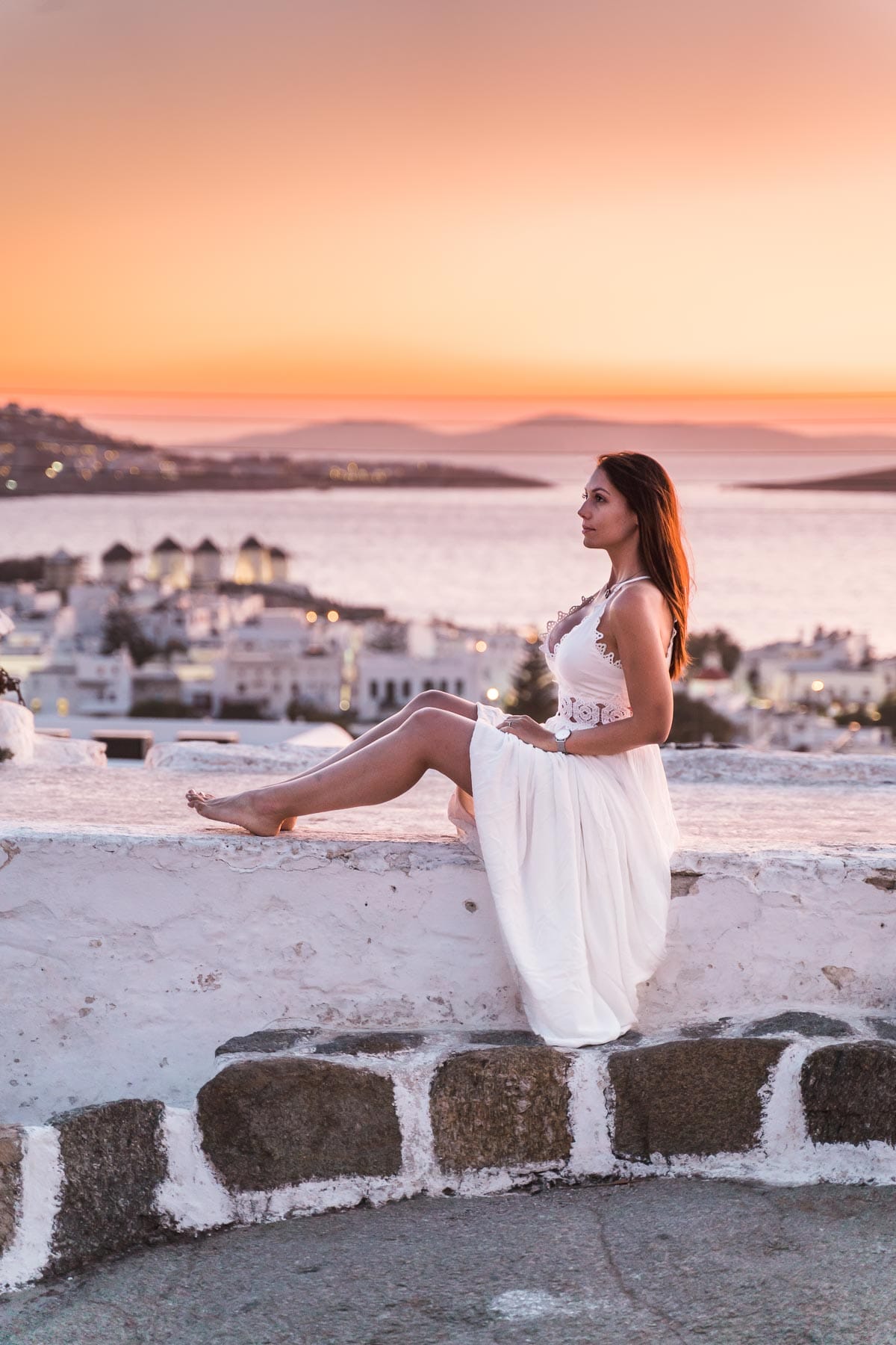 Girl in a white dress sitting on a wall at Boni's Windmill, watching the sunset over Mykonos Town