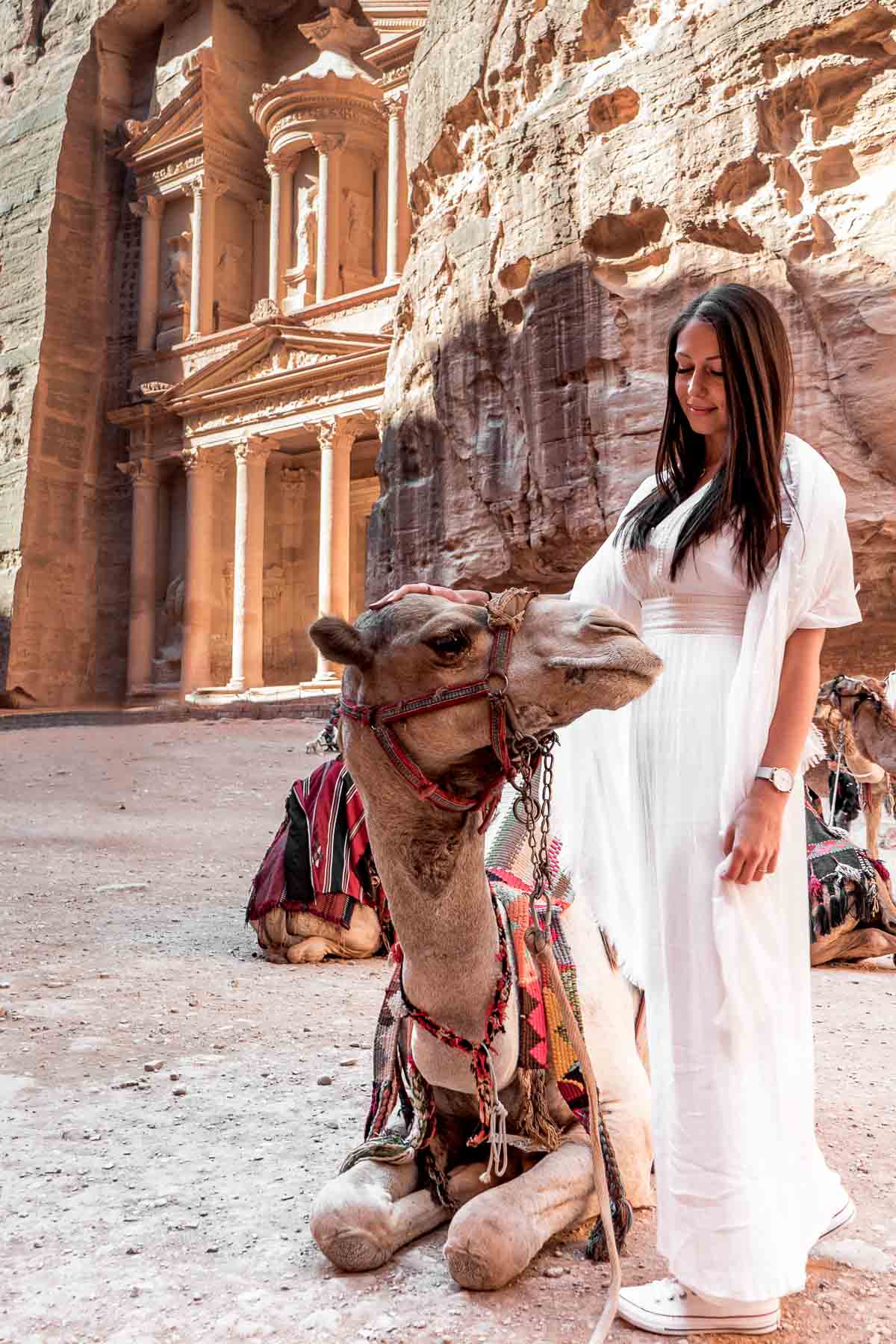 Girl in a white dress standing next to a camel with the Treasury in the background at Petra, Jordan