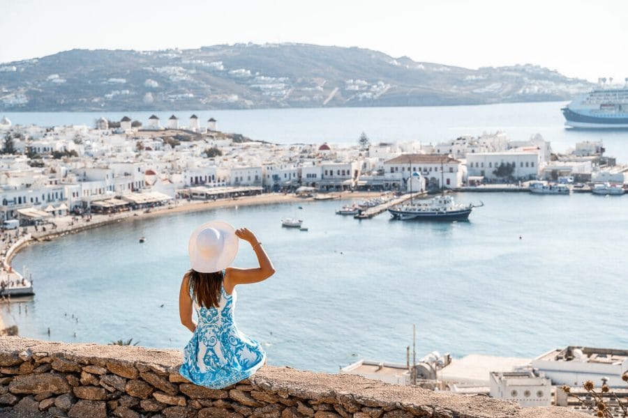 Girl in a blue dress sitting on a wall with the view of Mykonos Town in the background