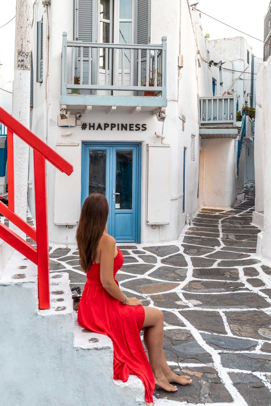 Girl in a red dress sitting on the stairs in front of the #happiness store which is one of the most instagrammable places in Mykonos