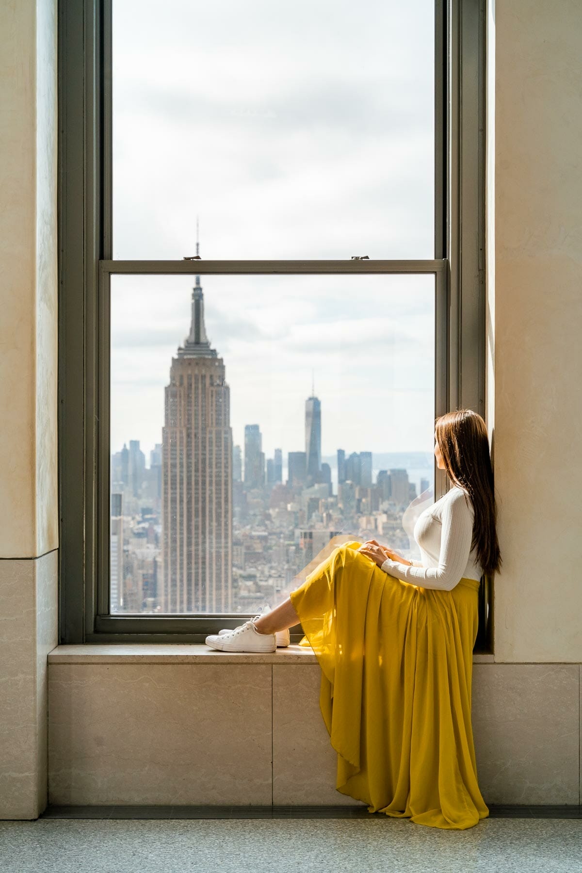 Girl in yellow skirt sitting in a window, overlooking the Empire State Building at Top of the Rock